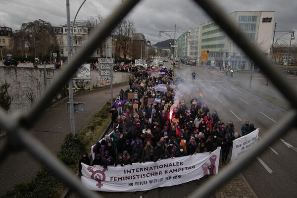 Durch ein Drahtzaunloch ist die Demofront der feministischen Kampftagdemo zu sehen, aus der Wilhelmstr. kommend fast vor der Blauen Brücke. Ein Bengalo scheint aus dem Block, in dem viele lila Halstücher tragen.