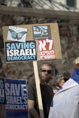 Eine Person hält ein Schild auf einer Holzleiste in die Höhe. Darauf steht u.a.: &quot;Saving isreli democracy&quot;.