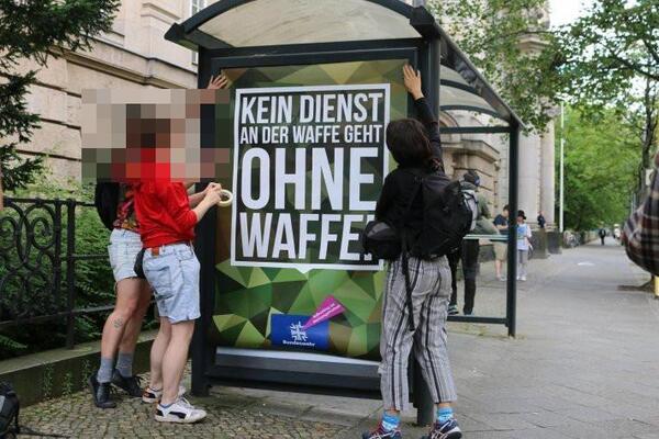 Eine Gruppe Menschen hängt ein Plakat in ein Bushäuschen im Camouflage-Stil der Bundeswehr: &quot;KEIN Diensta an der Waffe geht OHNE Waffe&quot;