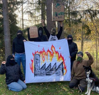Vermummte Aktivisti* mit Hund posieren um ein Transparent mit brennender Fabrik mit erhobenen Fäusten und auf Pappschildern geschriebenen Forderungen