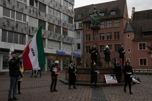 Auf dem Kartoffelmarkt stehen Menschen, von denen jeweils ein Auge mit blutigem Pflaster zugeklebt ist. Ein Mensch links hält eine Iran-Fahne, daneben ein Mensch mit Schildern in der Hand. Die weiteren haben Plastiklaschen als Rasseln in der Hand.