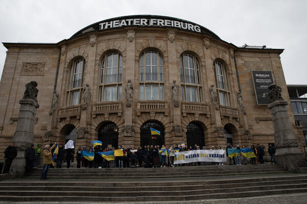 Blick auf das Freiburger Stadttheater. Vor den Toren stehen Demonstrierende aufgereiht mit einem großen Banner, auf dem &quot;We stand with Ukraine&quot; steht. Viele Fahnen der Ukraine sind zu sehen.