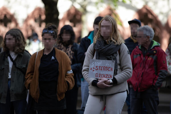 Mehrere Menschen stehen in Jacken gekleidet nebeneinander. Eine Person mit langen, blonden Haaren hält ein Schild vor ihren Bauch, auf dem steht: &quot;Stopt das Sterben an unseren Grenzen&quot;
