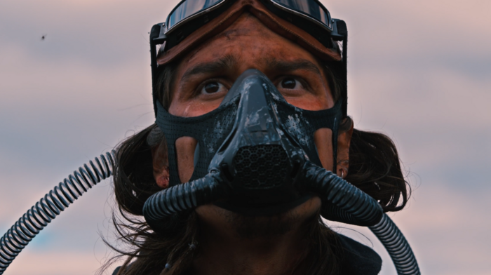 Ein junger Mann mit Gasmaske, verdrecktem Gesicht und hochgeschobener Fliegerbrille vor einem Himmel mit Schäfchenwolken, den Blick neben die Kamera in die Ferne gerichtet