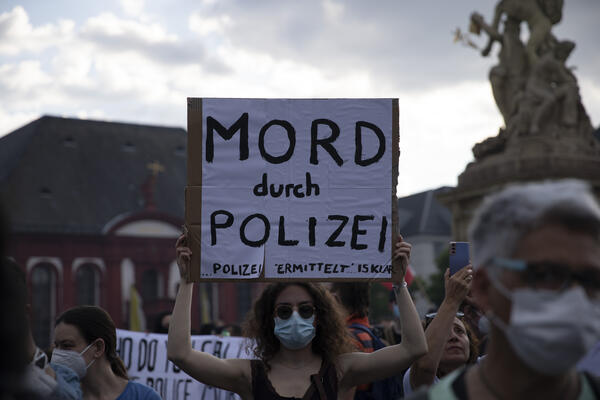 Eine weiblich gelesene Person mit langen, braunen Locken, Sonnenbrille und OP-Maske, hält ein Schild in die Höhe, auf dem steht: &quot;Mord durch Polizei - Polizei ermittelt, ist klar&quot;