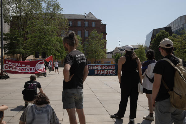 Menschen stehen auf dem Platz der Alten Synagoge und sind mit dem Rücken zur Kamera gerichtet. Hinten sind zwei Transparente zu sehen: Antifa in die Offensive, steht auf rotem Stoff. Gemeinsam kämpfen, auf dunkelblauem.
