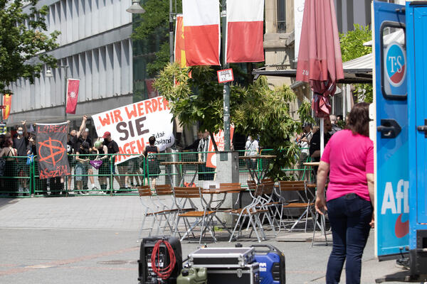 Im Vordergrund am rechten Bildrand das Heck eines Blauen AfD-Trucks, eine Person mit pinkem Shirt und Soundtechnik. Im Hintergrund in der linken Bildhälfte Antifaschist*innen. Manche zeigen den Stinkefinger, außerdem ist u.a. eine Anarchie-Fahne zu sehen.