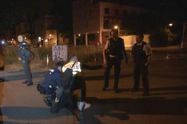 Pressesprecher der Polizei kniet auf einer dunkel gekleideten Person und zeigt mit dem rechten Arm in die rechte Richtung. Links daneben sitzt ein weiterer Polizist auf der Person. Rechts stehen zwei Beamte und schauen auf die Maßnahme.