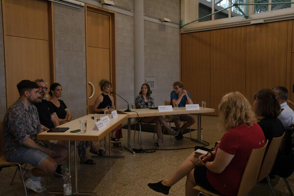 An zwei schräg zueinander gestellten Tischen sitzen von links nach rechts Simon Waldenspuhl, Stephan Schleith, Miriam Künzel, Hedra Youkhana, Anke Nöller und Julian Wiedermann. Am rechten Bildrand sind drei Menschen des Publikums zu sehen.