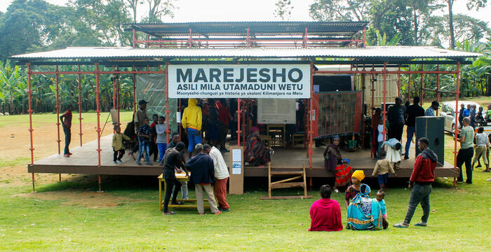 Ausstellung Marejesho am Kilimanjaro - ein Pavillon auf der Wiese mit vielen Besucher*innen. Im Hintergrund Bananenstauden.