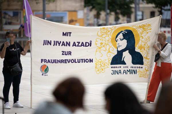 Zwei Personen halten ein Transparent, an dem Stöcke angebracht sind. Bild und Name von Jina Amini ist zu lesen, das Logo von TJK-E und &quot;Mit Jin Jiyan Azadi zur Frauenrevolution&quot; sind ebenfalls zu sehen.