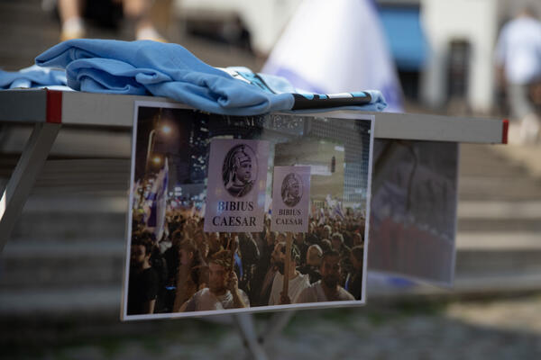 Nahaufnahme eines Fotos, das an einen Tisch geklebt ist. Darauf sind Protestierende mit Schildern zu sehen, die Netanjahu als &quot;Bibius Caesar&quot;, als römischen Diktator, darstellen.