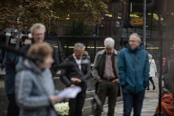 Im Vordergrund sind verschmonnene Menschen zu sehen, u.a. Irina Katz mit Blättern in der Hand vor einem Mikrofon. Im Hintergrund das Straßenschild &quot;Gurs&quot;, im Fokus.