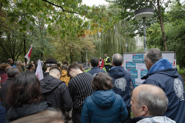 Menschenmenge der Uni Freiburg, zwei Menschen davon mit Jacken mit Uni-Logo, schauen auf zwei Redner*innen. Die eine Person trägt eine rote Verdi-Jacke, die andere eine Warnweste. Am rechten Bildrand ist eine Tafel mit Forderungen der Gewerkschaft zu sehn