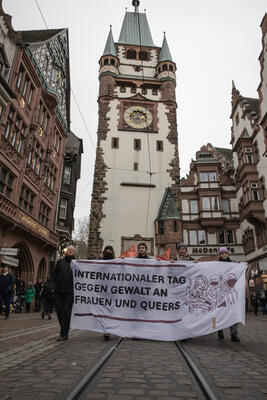 Hochformat. Demospitze mit weißem Transparent mit 5 Leuten, die es halten vor dem Martinstor: &quot;Internationaler Tag gegen Gewalt an Frauen und Queers&quot;