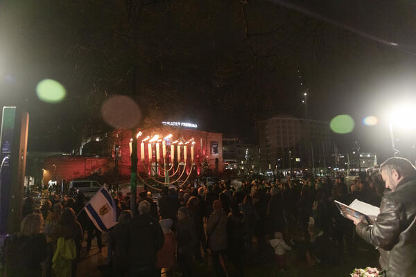 Auf dem Platz der Alten Synagoge blickt eine Menschenmenge auf eine Chanukkia. Es ist Dunkel. Von den Bildseiten strahlen zwei Lichter. Unten rechts steht Moshe Hayoun mit aufgeschlagenem Liedbuch.