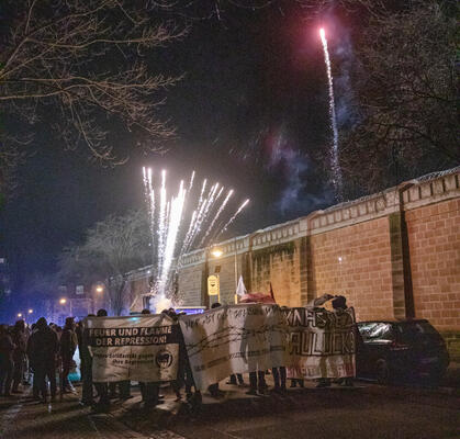 Eine Demonstration steht vor einer Gefängnismauer. Es ist Feuerwerk zu sehen und drei weiße Transparente.