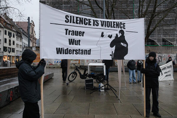 Zwei dunkel und winterlich gekleidete Menschen halten ein Banner mit zwei Holzlatten in die Höhe: Darauf abgebildet ein Polizist, der an einer schwarzen Person Gewalt ausübt. In schwarzen Druckbuchstaben steht: Silence is violence. Trauer, Wut, Widerstand