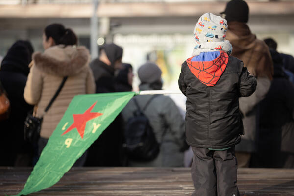 Ein kleines Kind ist von hinten zu sehen, es trägt eine helle Mütze und eine schwarze Jacke mit roter Kapuze. Es hält eine YPJ-Fahne auf der linken Seite.