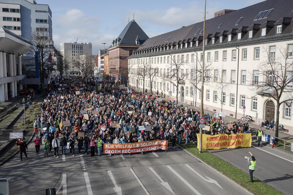 Ein riesen Demonstrationszug zieht sich über den kompletten Friedrichsring. Vorne ein Mensch in gelber Weste und Megafon streckt der Demo seine Faust entgegen. Zwei Fronttransparente, eines davon: Für Vielfalt &amp; Solidarität - Gegen Rechtsextremismus.