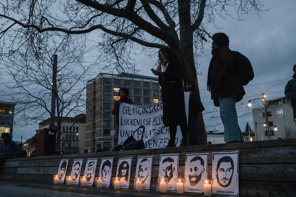 Zwei Menschen stehen auf einer Empore auf dem Platz der Alten Synagoge im Dunkeln. Unten sind neun Portraits der Ermordeten von Hanau zu sehen, davor jeweils eine Kerze.