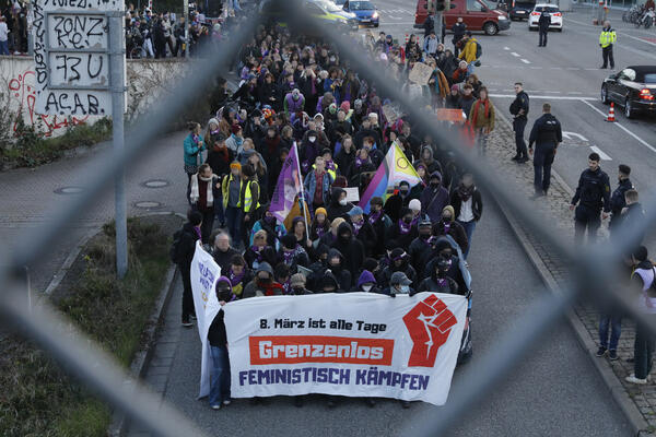 Demofront durch eine Metallzaunöffnung von oben fotografiert. Auf dem Transparent mit roter Faust steht: 8. März ist alle Tage - Grenzenlos - Feministisch Kämpfen.