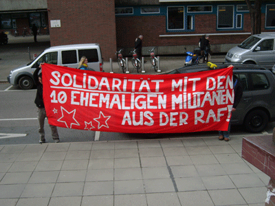 Demonstrierende auf einer Straße vor Hausfassade halten ein rotes Transpi auf dem steht: &quot;Solidarität mit den 10 ehemaligen Militanten aus der RAF!&quot;