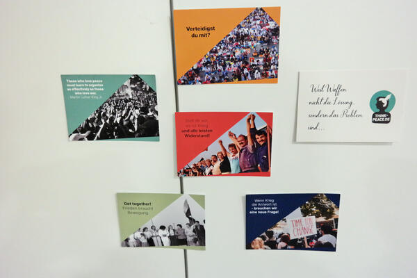 Wand mit Postkarten: U.a. Verteidigst du mit? Stell dir vor es ist Krieg und alle leisten Widerstand (Menschen nehmen sich an die Hand)