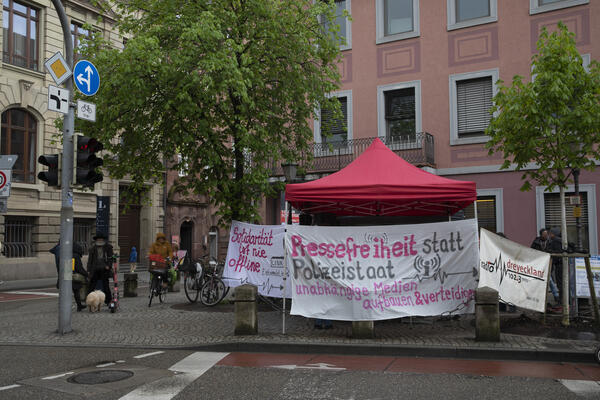 Ein roter Pavillion steht im Regen an der Straßenseite. Es sind verschiedene Transparente die zur Verteidigung der Pressefreiheit aufrufen angebracht und ein Banner von Radio Dreyeckland.