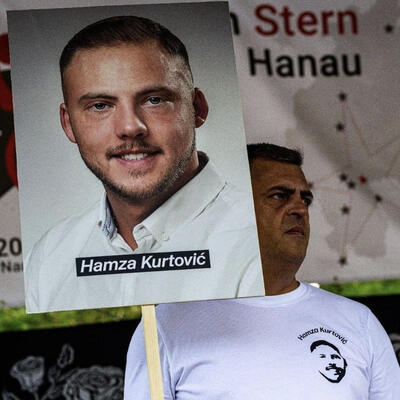 Armin Kurtović hält ein Schild mit dem Bild seines Sohnes Hamza Kurtović hoch. Er trägt ein weißes T-Shirt mit dem Konterfei seines ermordeten Sohnes. Im Hintergrund ist ein großes Banner zu sehen, ein Teil des Schriftzuges ist &quot;Hanau&quot;.