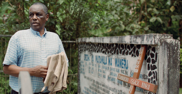 Eine Person steht neben einem Grabstein. Daran ist ein Holzkreuz gelehnt mit dem Namen Eliashukura Issack Nnko. Die Person trägt ein kariertes Hemd und hat einen Lappen in den Händen. Er schaut zur Seite, an der Kamera vorbei. Im Hintergrund stehen Bäume