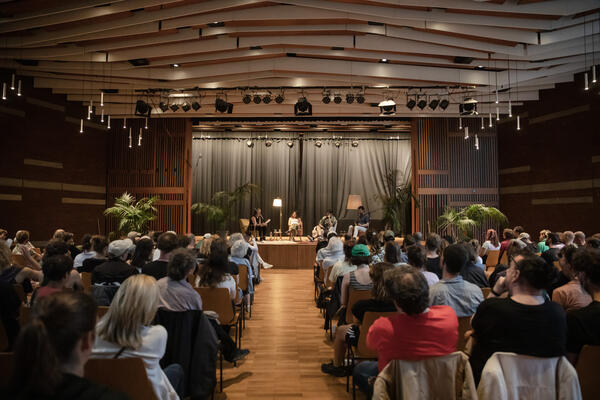 Blick von hinten in den Saal der Haus der Jugend. Viele Menschen sitzen auf den Stühlen. In der Mitte Gang frei, Parkettboden. Im Hintergrund im Fokus die Bühne mit vier Menschen darauf. Sie sitzen auf Sesseln und es gibt zwei Lampen und Zimmerpflanzen,
