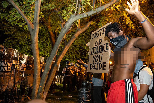 George Floyd Protest in Washington DC - Ein protestierender Mann mit schwarzem Tuch vor dem Mund. oberkörperfrei, mit einem Schild „Gerechtigkeit oder Gewalt, du entscheidest“. Ihm gegenüber stehen schwer ausgerüstete Militärpolizisten.