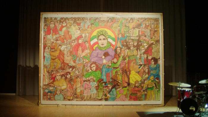 Ein buntes Gemälde auf einer Bühne, nebendran ein Schlagzeug. Das Bild, nach traditioneller iranischer Art gemalt, zeigt in der Mitte eine Frau mit grünem Kopftuch und Kind in der Armbeuge; dahinter die iranische Flagge. Drumherum teils bewaffnete Persone