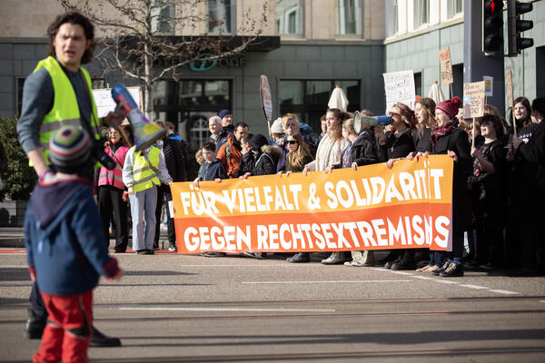 Gelb-orangenes Frontbanner mit &quot;Für Vielfalt &amp; Solidarität - Gegen Rechtsextremismus&quot; und Menschen im Hintergrund. Vorne links steht ein Mensch mit langen, lockigen Haaren, Megafon und gelber Warnweste. Davor ein Kind mit Mütze und Jacke.
