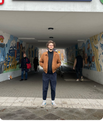 Niklas Schenker steht unter einer Unterführung mit Graffiti. Er trägt Turnschuhe und ein legeres Outfit mit Pulli und blauer Hose. Die Haare sind verwuschelt, die Hände in den Jackentaschen schaut er herausfordernd in die Kamera