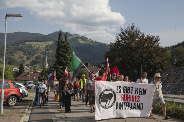 Auf einer schmalen Straße im Wohngebiet zieht eine Demonstration bergauf. Im Hintergrund Schwarzwald-Panorama, im Vordergrund rote und eine schwarz-grüne Fahne und Fronttransparent: &quot;Es gibt kein ruhiges Hinterland&quot; mit dem Logo der aufrufenden Gruppe.