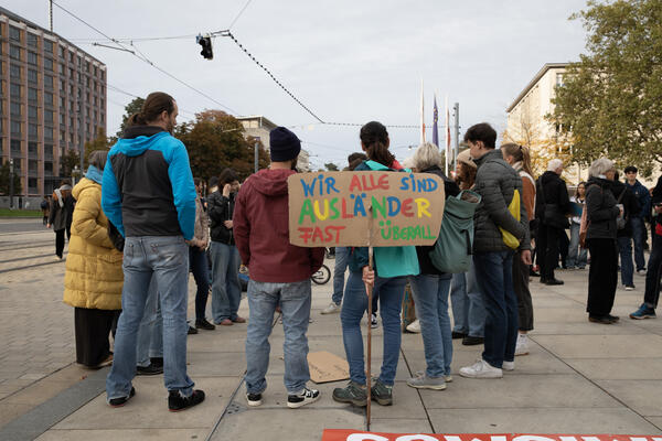 Eine Menschengruppe von ca. 14 Personen steht am Rande des Platz der Alten Synagoge am Rand. Im Hintergrund links Ausschnitt des Regierungspräsidiums. Rechts weitere Menschengruppen. Vorne buntes Schild: &quot;Wir sind alle Ausländer fast überall&quot;