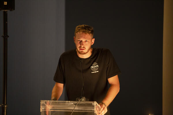 Omri Epstein- Mensch mit kurzen blonden Haaren und schwarzem T-Shirt mit NOVA-Logo, steht an einem Pult.