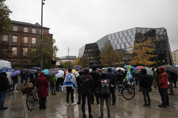 Menschen mit Schirmen und Regenjacken stehen mit dem Rücken zur Kamera auf dem Platz der Alten Synagoge. Im Hintergrund rechts ist die Uni-Bibliothek und links das KG1 der Uni zu sehen. Eine Person hat eine Israel-Fahne über den Rücken gehängt.