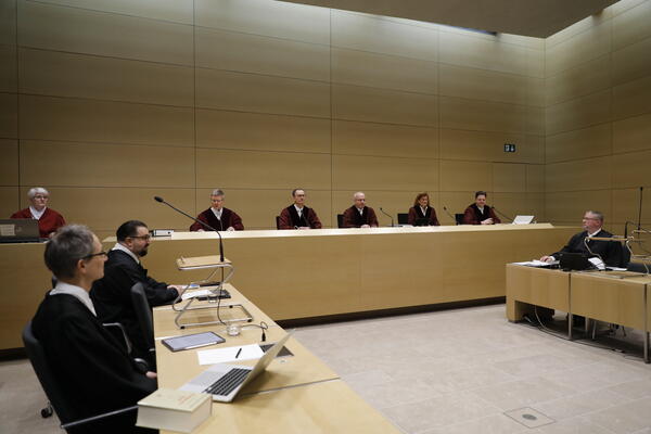 Blick in Richtung Verfahrensbeteiligte. Linker Bildrand zwei Verteidiger in schwarzen Roben. Hinten links die Protokollantin in roter Robe, daneben die Richterbank mit 5 Mitgliedern des Senat in schwarzen Roben. Rechter Bildrand, ein Neonazi-Anwalt.