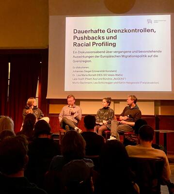 Auf dem Bild sind die Referent:innen abgebildet auf einem kleinen Podium, hinter ihnen das Bild des Beamers mit dem Namen der Veranstaltung. Viele Rücken der Zuhörenden Menschen ebenfalls.