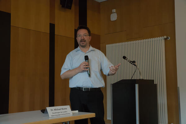 Dr. Michael Blume hält ein Mikrofon in der rechten Hand, mit der rechten gestikuliert er. Er steht aufrecht, neben einem Tisch, auf dem ein Namensschild mit seinem Namen zu sehen ist. Hinter ihm ein schwarzes Redepult mit weiterem Mikrofon.