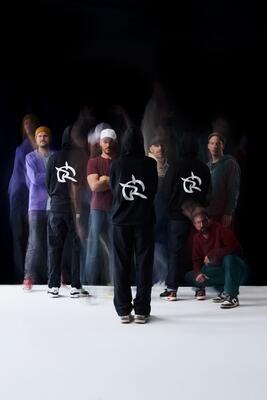 Irie Révoltés Pressefoto 2025 (c) Jamila K. Grote_2: Die neunköpfige Band teilweise mit schwarzen Kapuzenpullis und ihrem eigenen Logo vor dunklem Hintergrund. Einige Personen sind nur schemenhaft zu erkennen wie auf einem verwackelten Foto