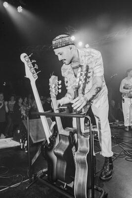 Ein Mann mit Kappe steht auf einer Bühne und greift zu einer Gitarre. Schwarz-weiß Foto.