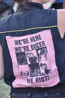 rosa Patch auf schwarzemGrund: den ganzen Rücken bedeckt das Patch einer Person in schwarz mit kurzen Ärmeln auf dem steht: We'r here We're queer We riot!