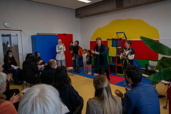 In einem Raum mit bunten Wänden stehen Menschen und schauen auf das Publikum. das mit dem Rücken zur Kamera links-unten im Bild zu sehen ist. Rechts sitzt Martin Horn. In der Bildmitte ist Sylvia Schliebe zu sehen, weitere Personen der Kita und Kinder.