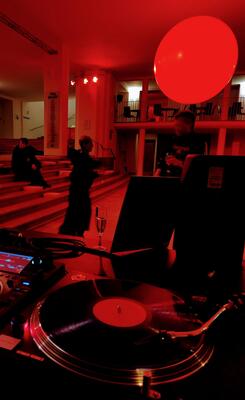 Im Vordergrund ist ein Plattenspieler zu sehen. Im Hintergrund zwei tanzende Menschen im Steinfoyer des Theaters.