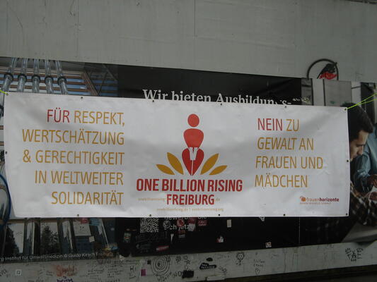 1 Banner zwischen 2 Bäumen. mittig steht rot auf weiß ONE BILLION RISING FREIBURG + Logo UND URL. Rechts und links steht in gelb: Für Restpekt, Wertschätzung &amp; Gerechtigkeit in weltweiter Solidarität. Nein zu Gewalt an Frauen und Mädchen. Frauenhorizonte