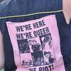 Ein Shirt auf dem ein Patch genäht ist. Darauf steht: We´re here, we´re queer, we riot 